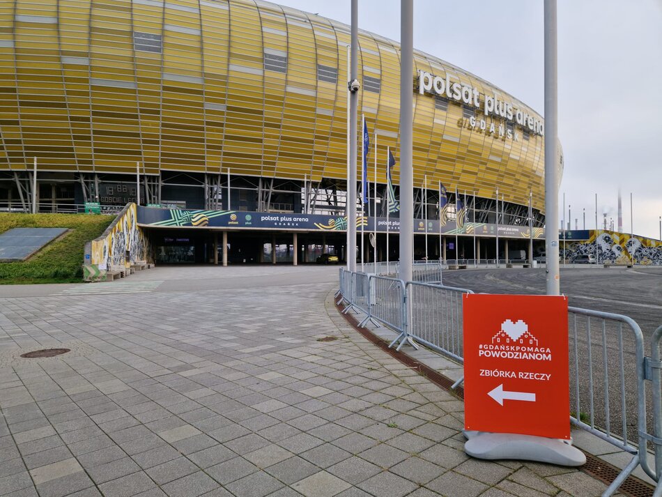 Zdjęcie przedstawia stadion Polsat Plus Arena Gdańsk z charakterystyczną złotą fasadą. Na pierwszym planie znajduje się czerwony znak informujący o zbiórce rzeczy dla powodzian, wskazujący kierunek za pomocą strzałki.