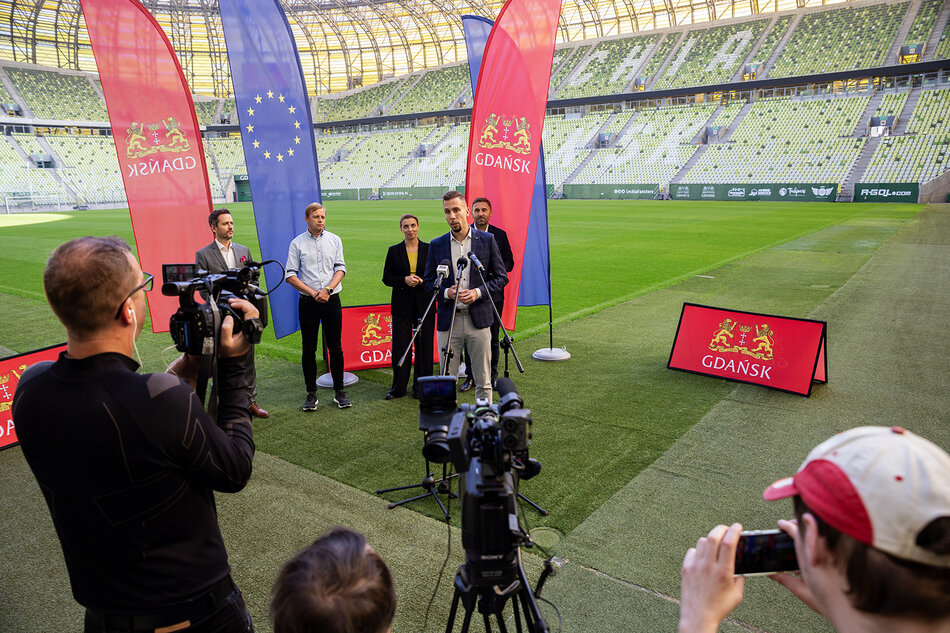 Konferencja prasowa na stadionie. Czterech mężczyzn i jedna kobieta stoją przed mikrofonami naprzeciw dziennikarzy. Za nimi murawa boiska.