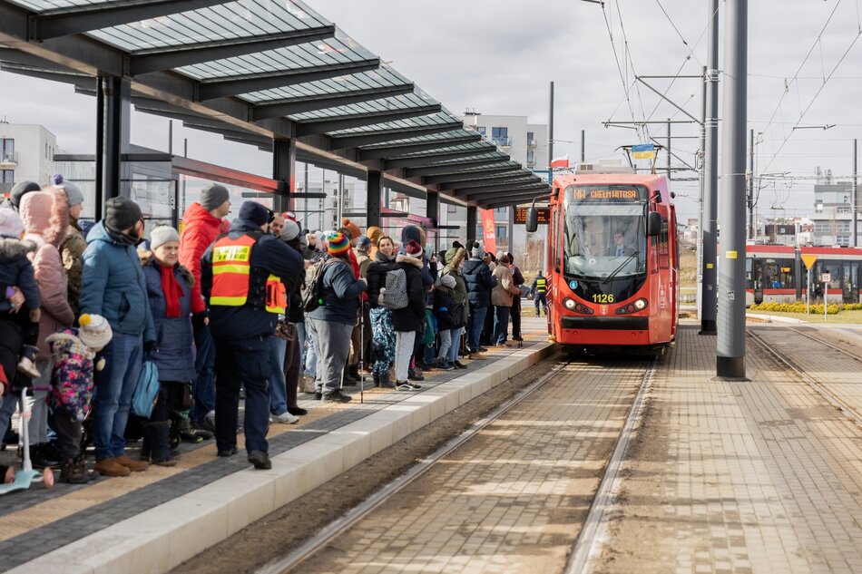 Na przystanku tramwajowym stoi tłum ludzi. Na przystanek podjeżdża tramwaj.