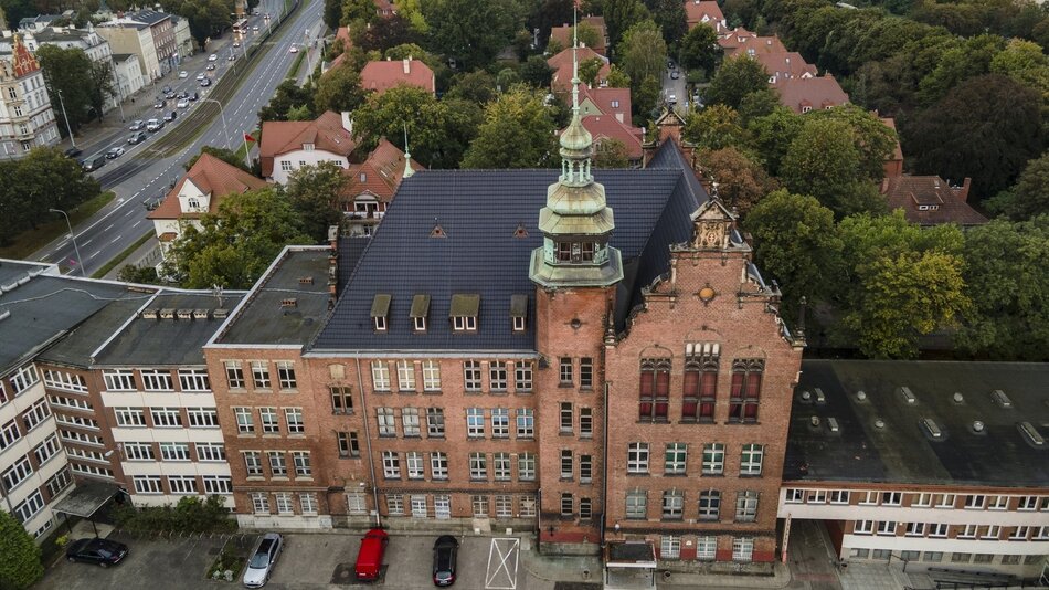 Na zdjęciu widać lotniczy widok dużego budynku z cegły o charakterystycznym stylu architektonicznym. Budynek posiada centralną wieżę zwieńczoną miedzianą kopułą. Architektura budynku jest klasyczna, z widocznymi łukowymi oknami i ozdobnym ceglanym detalem, co sugeruje, że może to być instytucja publiczna, np. szkoła lub urząd. Wokół budynku rozciągają się ulice i osiedla mieszkaniowe, z domami o czerwonych dachach, a także liczne drzewa, co świadczy o tym, że miejsce to znajduje się w zielonej, prawdopodobnie europejskiej okolicy. Obok budynku biegnie ulica z torami tramwajowymi oraz zaparkowanymi samochodami, co wskazuje na ruchliwy rejon z dostępem do komunikacji publicznej.