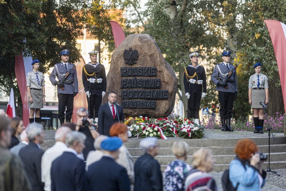 Zdjęcie przedstawia uroczystość upamiętniającą Polskie Państwo Podziemne, z żołnierzami i harcerzami stojącymi na straży przy pomniku, który ma formę kamienia z wyrytym napisem „Polskie Państwo Podziemne 1939-1945”. W tle widoczna jest zgromadzona publiczność, a na pomniku złożono wieńce i kwiaty, co nadaje uroczystości poważny, patriotyczny charakter