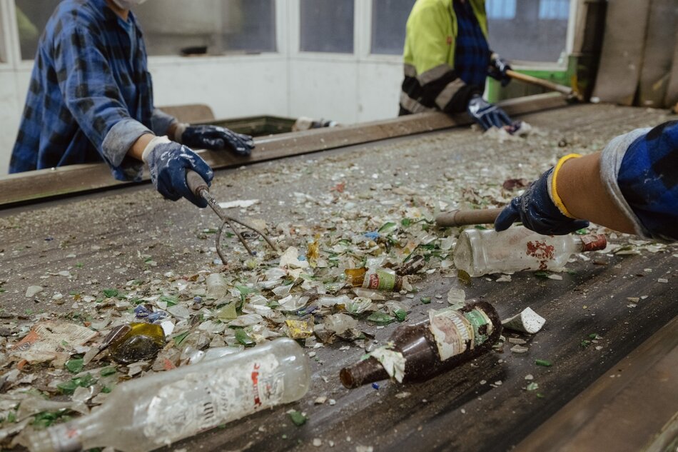 Na zdjęciu widzimy pracowników sortujących rozbite szkło na taśmie produkcyjnej, prawdopodobnie w zakładzie recyklingu. Noszą oni rękawice ochronne i korzystają z narzędzi do przemieszczania butelek i odłamków, co wskazuje na proces ręcznej segregacji szkła.