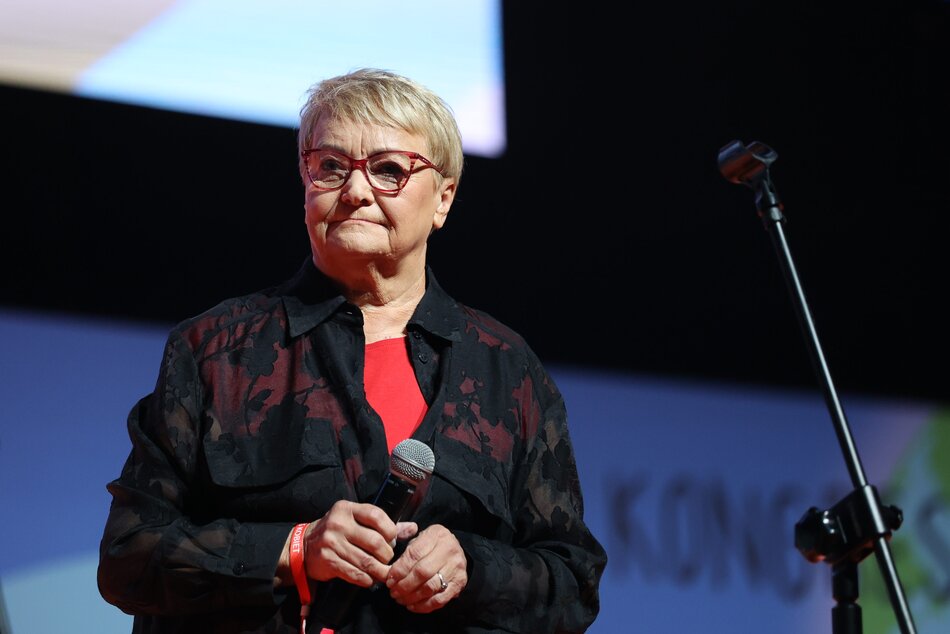 Na zdjęciu widzimy starszą kobietę w ciemnej bluzce, trzymającą mikrofon, stojącą na scenie. Wygląda na to, że uczestniczy w oficjalnym wydarzeniu, być może przemawia lub przygotowuje się do wystąpienia.