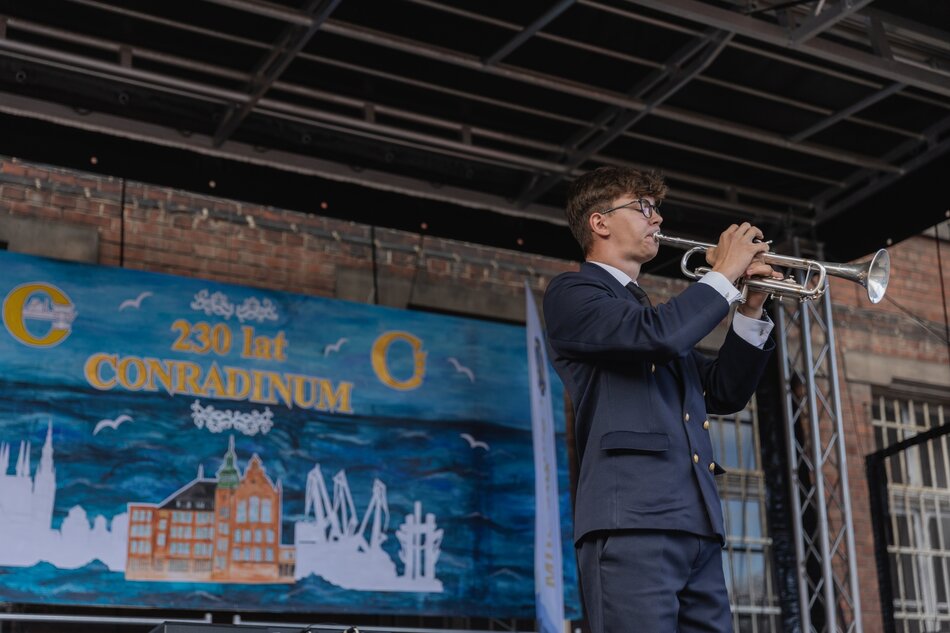 Na zdjęciu widzimy młodego mężczyznę w eleganckim mundurze grającego na trąbce na scenie plenerowej. W tle znajduje się baner z napisem 230 lat Conradinum, co sugeruje, że jest to uroczystość jubileuszowa związana z tą instytucją.