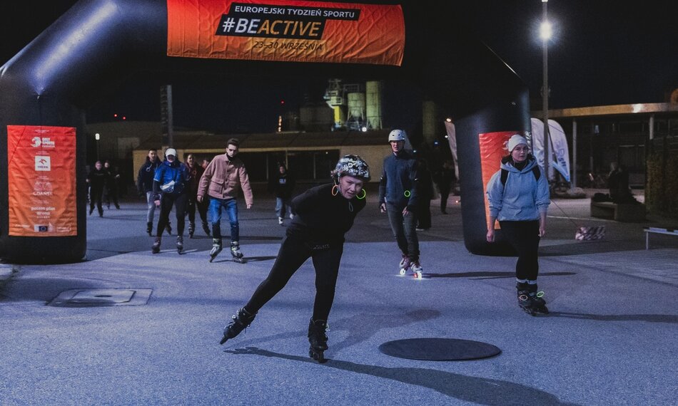 Na zdjęciu widzimy grupę osób jeżdżących na rolkach, które biorą udział w wieczornej imprezie sportowej pod hasłem BeActive. W tle widoczny jest duży nadmuchiwany łuk z napisem Europejski Tydzień Sportu oraz datami wydarzenia (23-30 września).