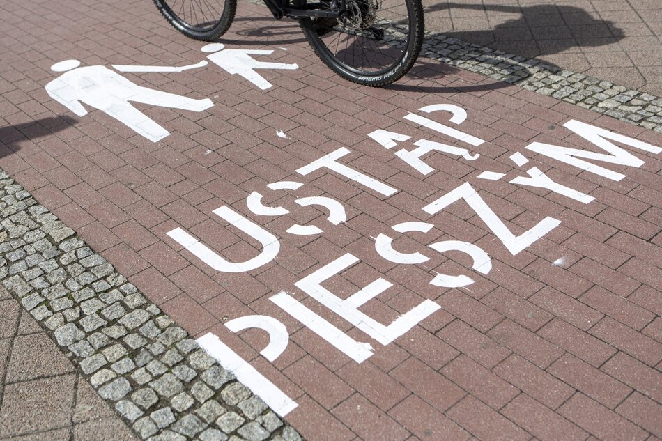 Na zdjęciu widoczna jest ścieżka rowerowa z namalowanym białym napisem USTĄP PIESZYM oraz symbolami pieszych, sugerującymi pierwszeństwo dla pieszych. Częściowo widoczny jest również rower, który znajduje się nad napisem, co wskazuje, że zdjęcie zrobiono w miejscu wspólnego ruchu rowerowego i pieszego.