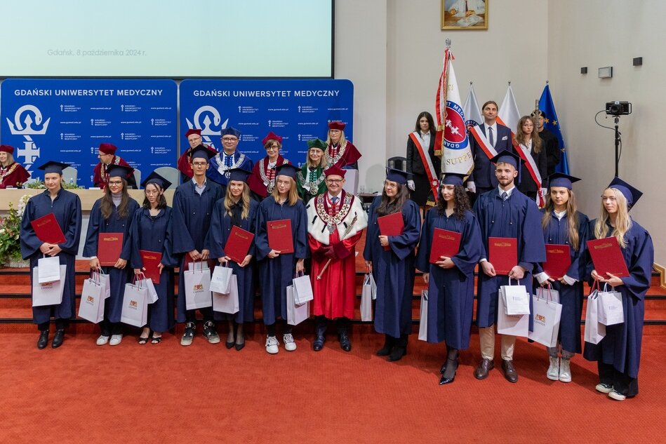 Na zdjęciu widzimy grupę absolwentów Gdańskiego Uniwersytetu Medycznego ubranych w togi i birety, trzymających dyplomy i torby upominkowe. W tle znajdują się przedstawiciele uczelni w oficjalnych strojach oraz sztandar uniwersytetu, co sugeruje, że zdjęcie zostało zrobione podczas ceremonii ukończenia studiów lub immatrykulacji