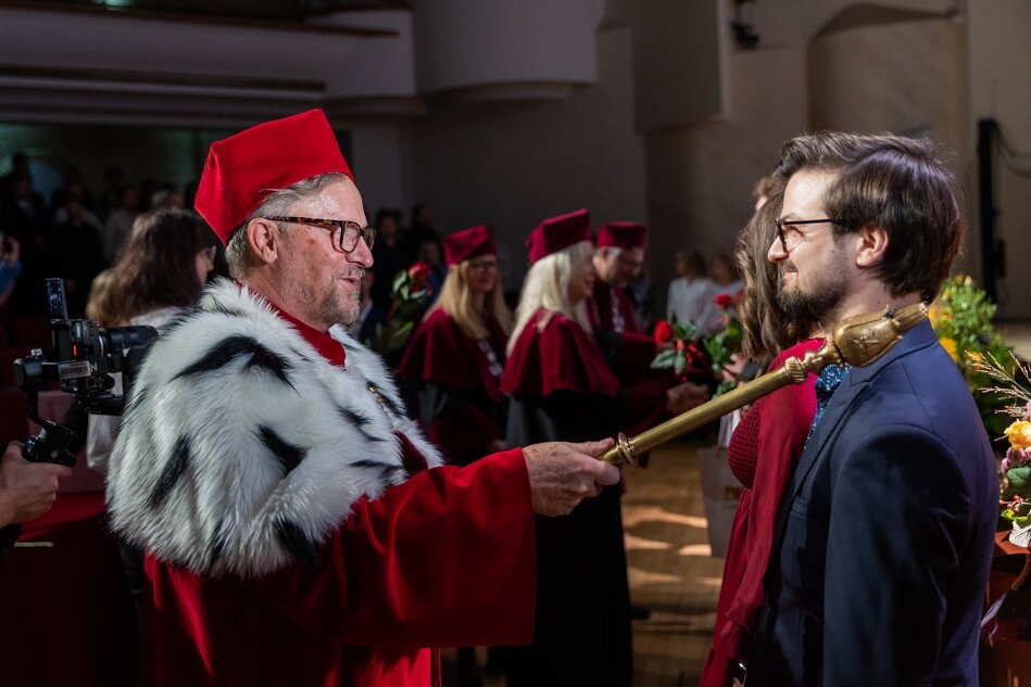 Na zdjęciu widać mężczyznę w tradycyjnym stroju akademickim - rektora, który trzyma ceremonialną laskę i wręcza ją młodemu mężczyźnie ubranym w garnitur. W tle można dostrzec inne osoby w togach. Scena odbywa się podczas immatrykulacji 