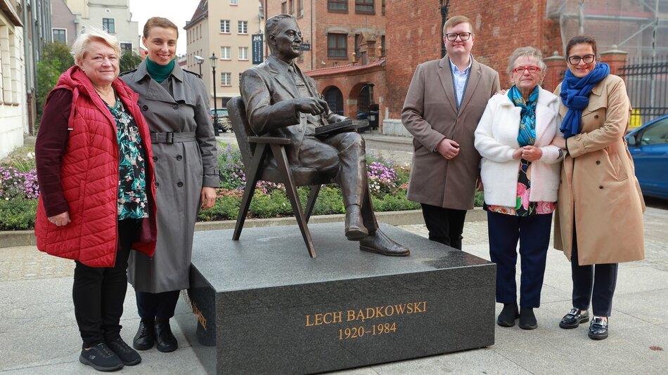 Na zdjęciu znajduje się grupa pięciu osób pozujących obok pomnika Lecha Bądkowskiego. Pomnik przedstawia postać Lecha Bądkowskiego siedzącą na krześle, z jedną nogą założoną na drugą. Na cokole pomnika widnieje napis „Lech Bądkowski 1920–1984”. Osoby na zdjęciu stoją w rzędzie obok pomnika, uśmiechają się i są ciepło ubrane. W tle widać miejską scenerię, z budynkami, roślinami oraz brukowaną ulicą.