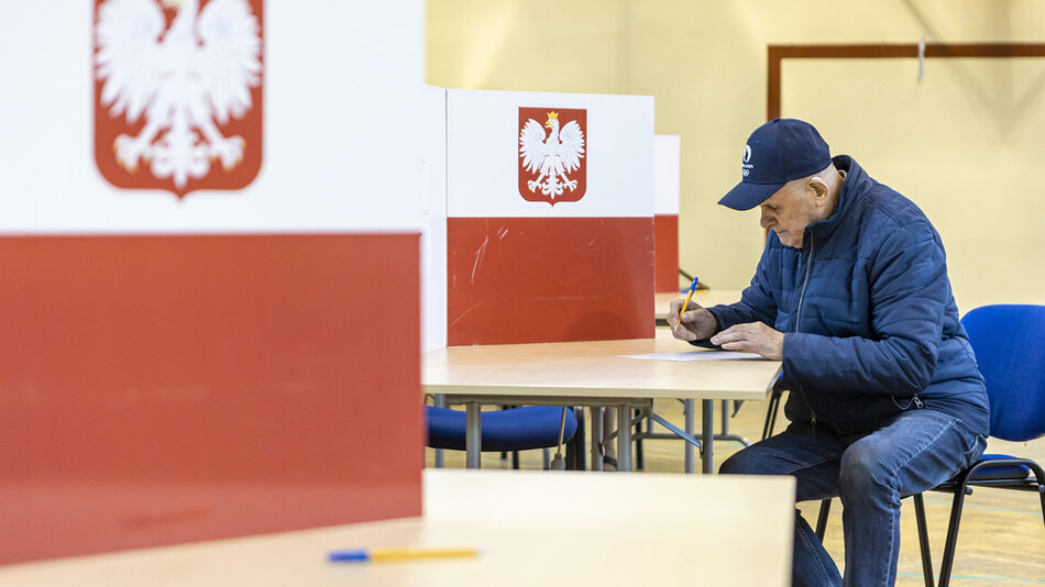 starszy mężczyzna w czapce z daszkiem i kurtce pisze coś na kartce, przy stoliku, we wnętrzu sportowej sali. Mężczyzna jest otoczony parawanami pomalowanymi w biało-czerwone barwy z godłem w kształcie orła na środku