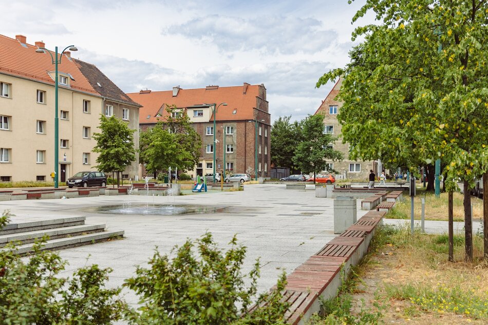 na zdjęciu osiedlowy plac z kostki chodnikowej, w tle kilka kamienic z czerwonymi dachami