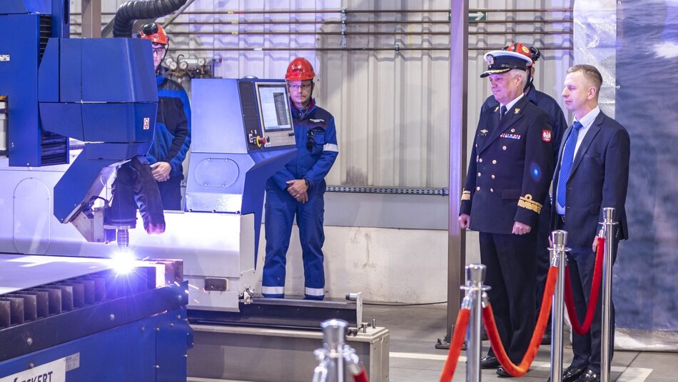 Na zdjęciu widać moment uroczystego rozpoczęcia cięcia stali, prawdopodobnie w stoczni lub innym zakładzie produkcyjnym. Po lewej stronie widoczna jest nowoczesna maszyna do cięcia, z której wydobywa się intensywne światło, najpewniej od łuku plazmowego używanego do cięcia metalu. Obok maszyny stoi dwóch mężczyzn w niebieskich strojach roboczych i czerwonych kaskach, którzy prawdopodobnie obsługują urządzenie. Po prawej stronie zdjęcia znajdują się dwaj mężczyźni w garniturach, z których jeden jest wojskowym w mundurze marynarki wojennej, co sugeruje, że może to być związane z budową lub modernizacją okrętu wojennego. Obszar wokół nich jest oddzielony czerwoną taśmą, co podkreśla uroczysty charakter tego wydarzenia. Zdjęcie pokazuje moment technologiczny, połączony z oficjalną obecnością przedstawicieli marynarki i zarządu stoczni lub firmy odpowiedzialnej za ten projekt.