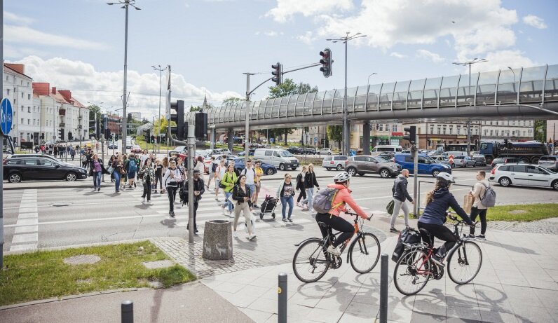 Na zdjęciu widzimy ruchliwe skrzyżowanie z dużą liczbą pieszych, rowerzystów i samochodów, które współdzielą przestrzeń. W tle znajduje się estakada oraz budynki, a cała scena rozgrywa się w mieście podczas pogodnego dnia, z niebem pokrytym częściowo chmurami.