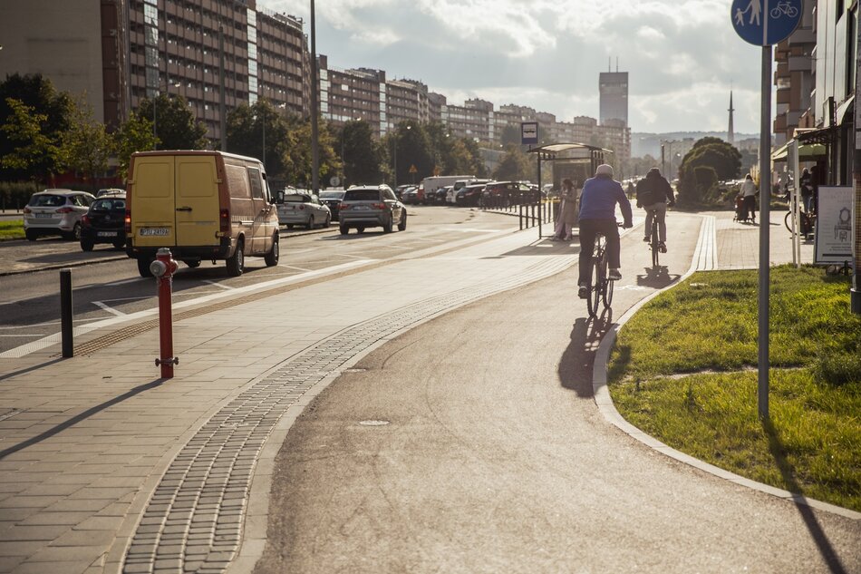 na zdjęciu ulica, widać drogę rowerową a na niej rowerzysta, obok chodnik, w tle po lewej jezdnia, a za nią wysoki i szeroki na kilkadziesiąt metrów budynek mieszkalny