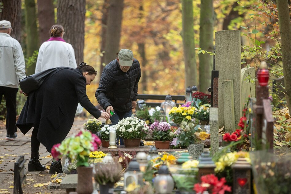 na zdjęciu kobieta i mężczyzna pochylają się nad kamiennym grobem, na cmentarzu, widać też inne groby