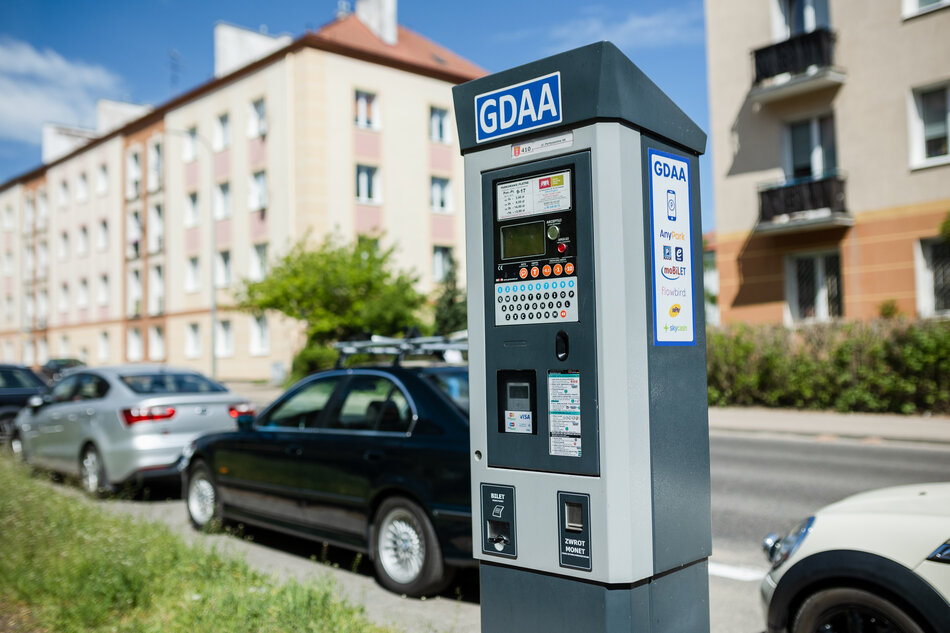 Na zdjęciu widać parkomat z oznaczeniem GDAA, znajdujący się przy ulicy z zaparkowanymi samochodami na tle budynków mieszkalnych. Parkomat umożliwia płatności za pomocą różnych aplikacji, takich jak AnyPark i moBILET.