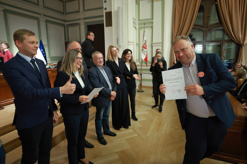 na zdjęciu po prawej mężczyzna w średnim wieku w garniturze pozuje do zdjęcia z dyplomem, po lewej stoi kilka osób w rzędzie, także elegancko ubranych