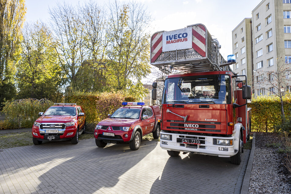 Trzy samochody straży pożarnej stoją obok siebie: dwa typu pick-up i jeden duży z drabiną. W tle zieleń