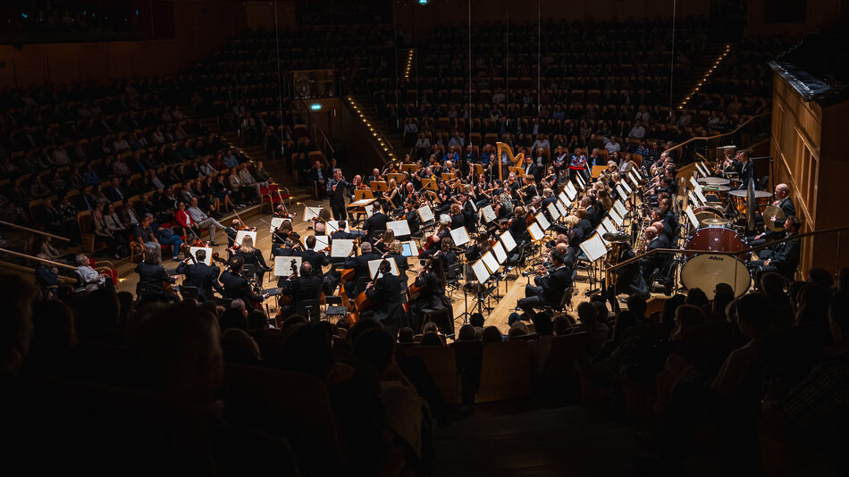 Na zdjęciu widoczna jest pełna orkiestra symfoniczna wykonująca koncert w dużej sali koncertowej, w której zgromadziła się licznie publiczność