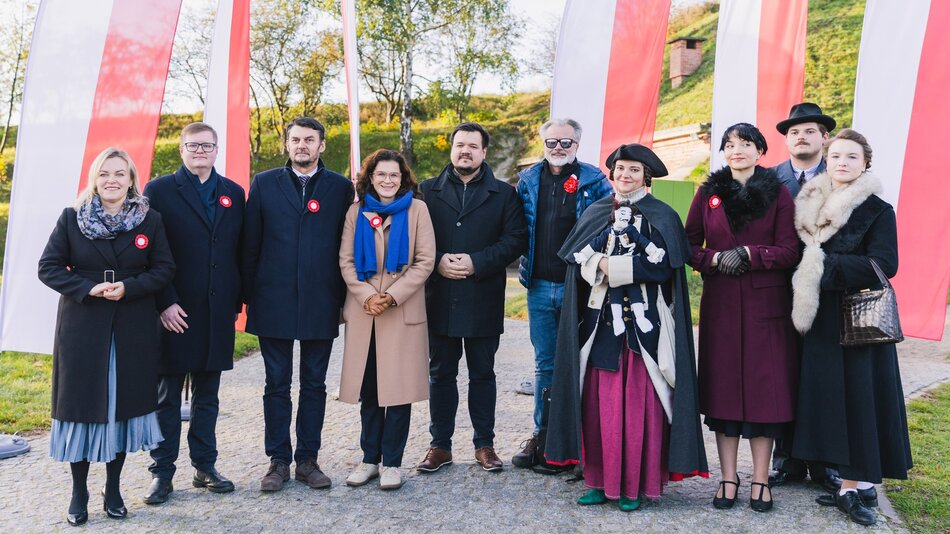 Na zdjęciu widzimy grupę osób pozujących na tle biało-czerwonych flag, prawdopodobnie z okazji narodowego święta w Polsce. Osoby są ubrane elegancko, niektóre z nich mają przypięte biało-czerwone kokardy – symbole patriotyczne. Część osób ubrana jest w współczesne stroje, jednak po prawej stronie zdjęcia znajduje się kilka osób w stylizowanych, historycznych ubiorach, co może sugerować, że w wydarzeniu brały udział osoby odgrywające rolę postaci historycznych lub rekonstruktorzy. Jedna z osób trzyma lalkę w historycznym stroju. Sceneria jest plenerowa, w tle widać zieleń i lekko pochmurne niebo.