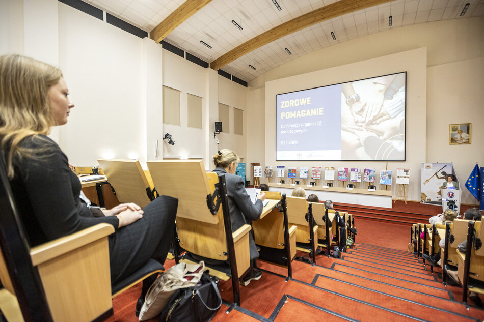 Na zdjęciu widzimy uczestników konferencji siedzących w auli, w której na dużym ekranie wyświetlany jest temat wydarzenia: Zdrowe Pomaganie – konferencja organizacji pozarządowych z datą 6.11.2024. W tle widoczne są plakaty i banery, a po prawej stronie mówca prowadzi prezentację przy mównicy, tworząc formalną atmosferę spotkania.