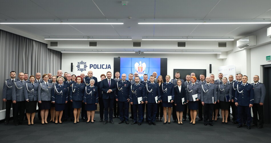 Na zdjęciu widzimy grupę funkcjonariuszy polskiej policji i pracowników administracji w eleganckich mundurach, zgromadzonych w sali konferencyjnej. W centrum znajduje się logo polskiej policji, a funkcjonariusze stoją równo w rzędach, niektórzy trzymają dokumenty, co sugeruje wręczenie nagród lub wyróżnień.