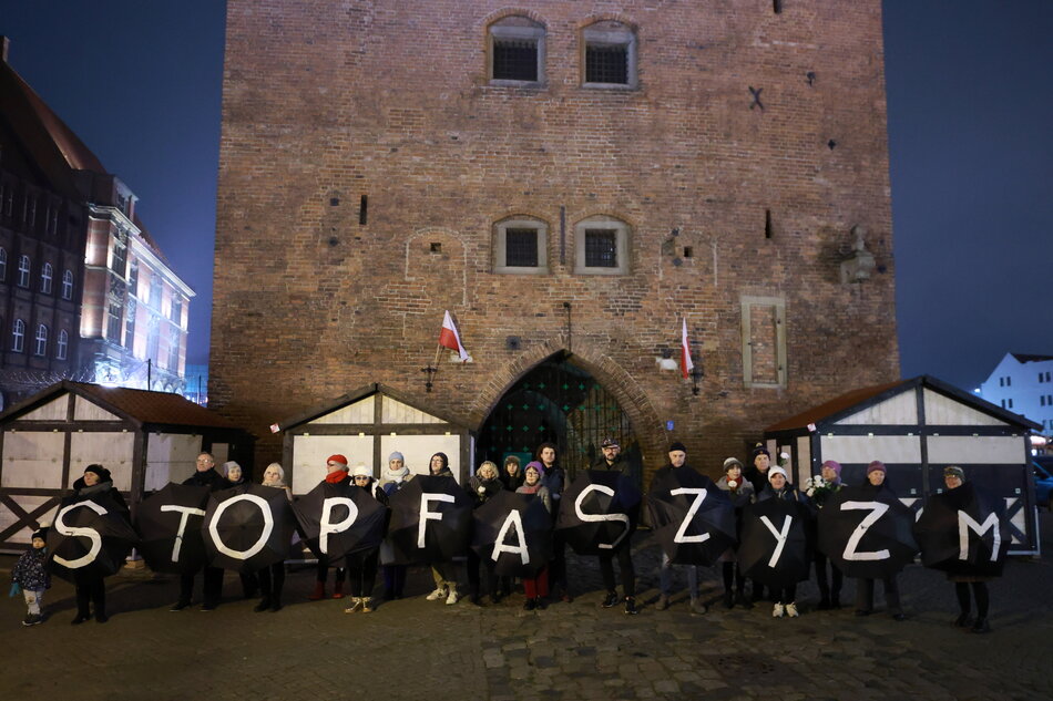 Grupa osób stoi pod średniowieczną wieżą i trzyma rozłożone czarne parasole, które tworzą napis stop faszyzm.