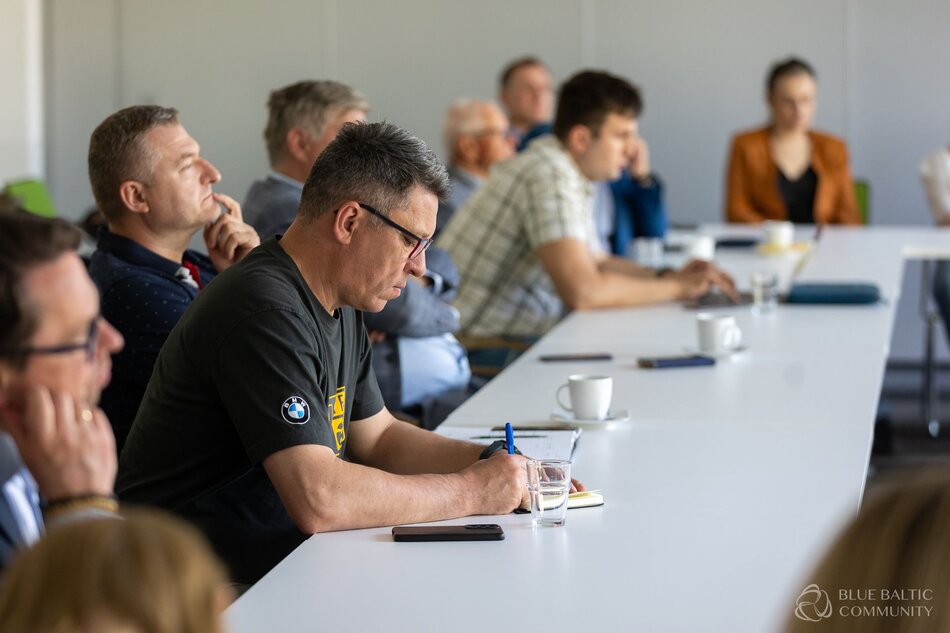 Zdjęcie przedstawia grupę ludzi siedzących przy długim stole konferencyjnym, skupionych na spotkaniu lub prezentacji. Osoby są uważne i skoncentrowane, niektórzy robią notatki, podczas gdy inni słuchają z zamyśleniem.