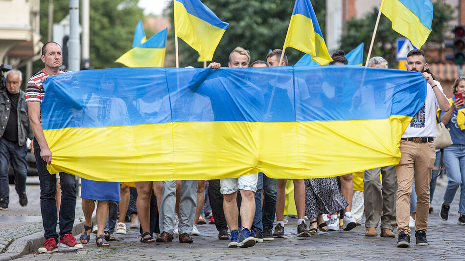 grupa ludzi idzie środkiem ulicy niosąc przed sobą dwu kolorową flagę. w tle widoczna grupa ludzi niosąca flagi w tych samych kolorach