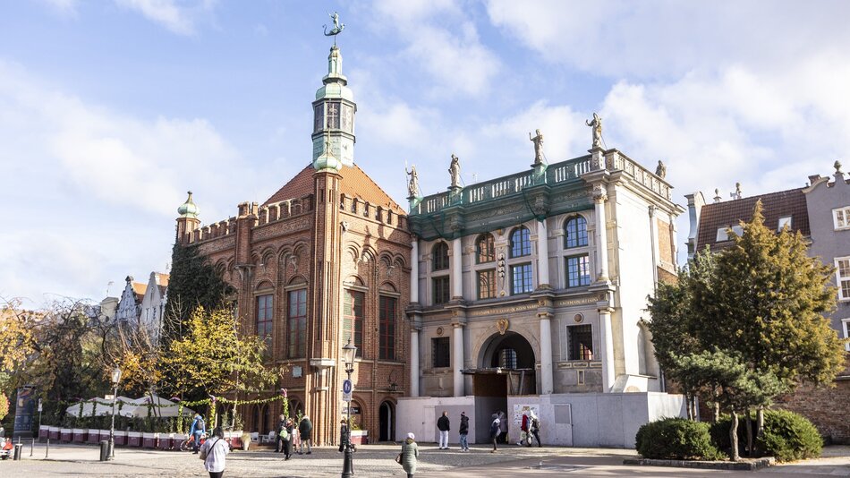 Na zdjęciu znajduje się fragment zabytkowej zabudowy Gdańska, przedstawiający dwa charakterystyczne budynki. Po lewej stronie widoczna jest Dwór Bractwa św. Jerzego, ceglany budynek w stylu gotyckim z charakterystycznymi łukami, ozdobnymi oknami i wieżyczką zwieńczoną kopułą oraz chorągiewką w kształcie smoka. Po prawej stronie widać Złotą Bramę, renesansowy budynek z jasną elewacją, kolumnami oraz bogato zdobionym gzymsem. Na balustradzie znajdują się rzeźby przedstawiające alegoryczne postacie. Na pierwszym planie widoczni są spacerujący ludzie, a także drzewa i ogródki restauracyjne ozdobione roślinnością. Całość sceny rozgrywa się w pogodny dzień z niebieskim niebem i rozproszonymi chmurami.