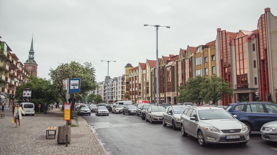 Na zdjęciu widzimy miejską ulicę w Europie, prawdopodobnie w Polsce, ze względu na oznaczenia drogowe i tablice rejestracyjne samochodów. Po prawej stronie znajdują się charakterystyczne, nowoczesne budynki z elementami w stylu postmodernistycznym, wykończone w jasnych kolorach z czerwonymi i złotymi akcentami. W tle widać wieżę kościoła z zielonym, spiczastym dachem. Po lewej stronie znajdują się starsze bloki mieszkalne z balkonami. Wzdłuż ulicy zaparkowanych jest wiele samochodów, a ruch na drodze wygląda na umiarkowany. Na chodniku po lewej stronie idzie kilka osób, a obok stoi przystanek autobusowy z rozkładem jazdy. Pogoda jest pochmurna, co sugeruje, że zdjęcie wykonano w chłodniejszym okresie roku. Atmosfera miejsca wydaje się spokojna i miejska.