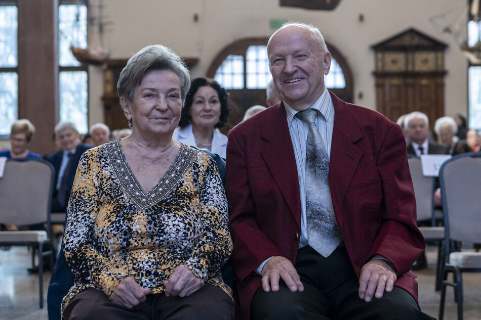 Na zdjęciu widoczna jest para seniorów siedzących obok siebie, uśmiechniętych, w eleganckim otoczeniu podczas uroczystości jubileuszowej. W tle widać innych uczestników wydarzenia oraz fragment stylowego wnętrza z dużymi oknami i zabytkowymi elementami