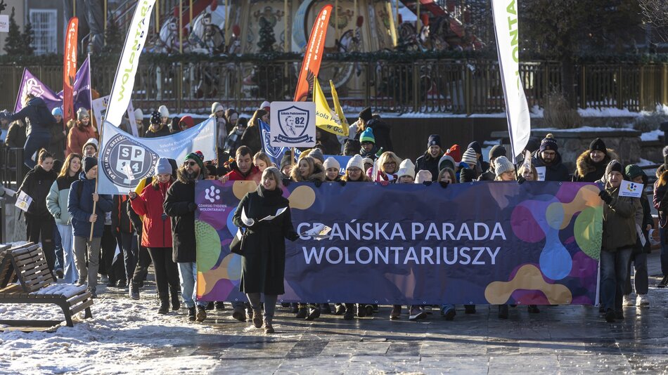 Na zdjęciu widoczna jest Gdańska Parada Wolontariuszy, która odbywa się w zimowej scenerii. Grupa uczestników maszeruje ulicą, trzymając duży, kolorowy baner z napisem „Gdańska Parada Wolontariuszy” oraz logotypem Gdańskiego Tygodnia Wolontariatu. W tłumie widać ludzi różnych grup wiekowych, ubranych w ciepłe zimowe kurtki i czapki, a także transparenty i flagi reprezentujące różne organizacje oraz szkoły, np. Szkołę Podstawową nr 82 czy SP 4. W tle można dostrzec karuzelę i inne świąteczne dekoracje, co sugeruje, że wydarzenie odbywa się w okresie zimowym, prawdopodobnie w grudniu. Atmosfera jest radosna i pełna zaangażowania, podkreślając wspólnotowy charakter wydarzenia oraz zaangażowanie w działalność wolontariacką. Na ziemi leży śnieg, co dodatkowo buduje zimowy nastrój.