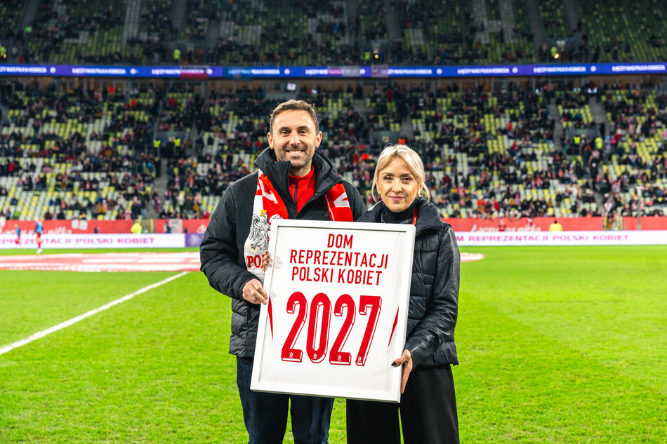 Na zdjęciu znajduje się dwoje ludzi na tle stadionu piłkarskiego. Trzymają tablicę z napisem Dom Reprezentacji Polski Kobiet 2027, co oznacza, że to miejsce będzie związane z kobiecą reprezentacją Polski w piłce nożnej do 2027 roku.