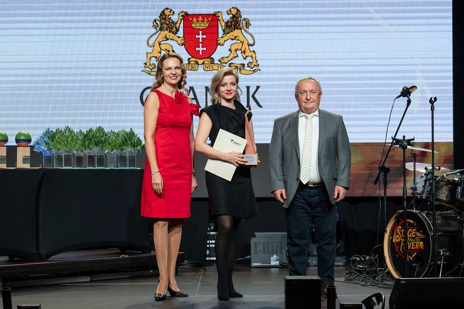 Na zdjęciu znajduje się troje osób pozujących na scenie podczas uroczystego wydarzenia. Pierwsza osoba z lewej strony to kobieta ubrana w elegancką, czerwoną sukienkę bez rękawów. Stoi uśmiechnięta, z rękami wzdłuż ciała. Druga osoba w środku to kobieta w czarnej sukience, trzymająca w dłoniach dokument lub dyplom. Wygląda oficjalnie i jest uśmiechnięta. Trzecia osoba z prawej strony to mężczyzna w szarym garniturze, białej koszuli i krawacie w delikatny wzór. Wygląda poważnie, stoi prosto. W tle widnieje herb miasta Gdańska oraz napis „Gdańsk”. Na stole z lewej strony znajdują się nagrody lub statuetki, ozdobione zielonymi elementami przypominającymi rośliny. Po prawej stronie widoczna jest perkusja i mikrofony, co sugeruje, że jest to gala z elementami muzycznymi.