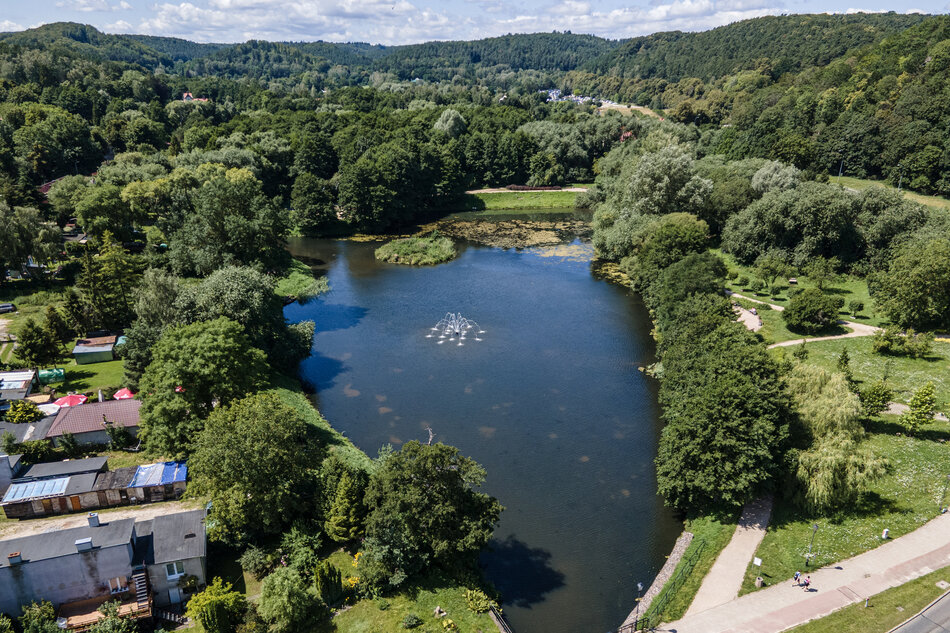 Zdjęcie przedstawia widok z lotu ptaka na malowniczy staw otoczony gęstą zielenią, z fontanną umieszczoną centralnie w wodzie. Wokół stawu widać ścieżki spacerowe oraz zabudowania i ogródki, tworzące spokojny i rekreacyjny krajobraz.