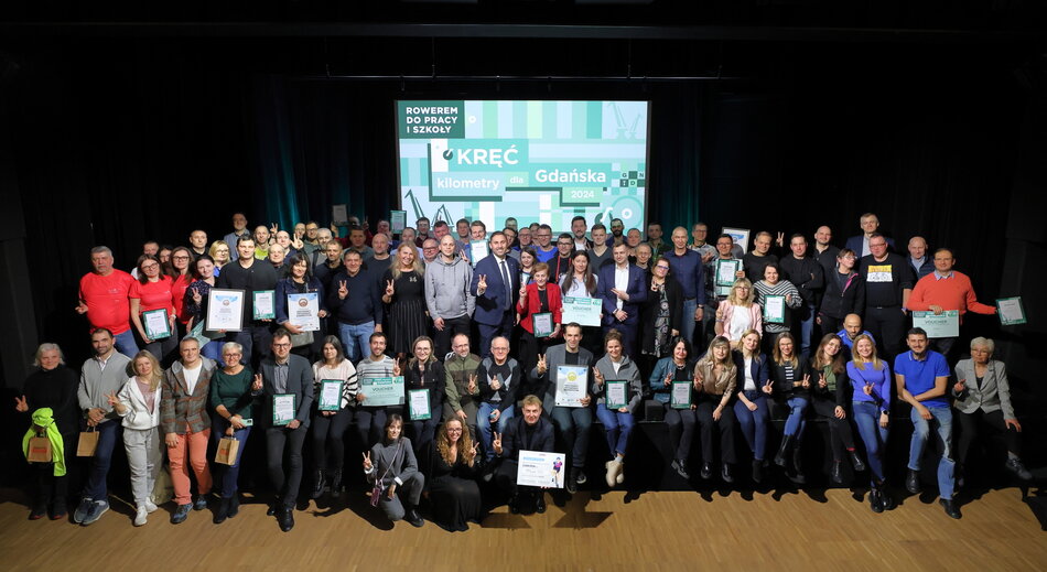 Na zdjęciu znajduje się duża grupa osób uczestniczących w wydarzeniu promującym rowerową aktywność w Gdańsku w ramach akcji Kręć kilometry dla Gdańska 2024. Uczestnicy pozują z dyplomami, voucherami i nagrodami, celebrując osiągnięcia w rowerowej rywalizacji.