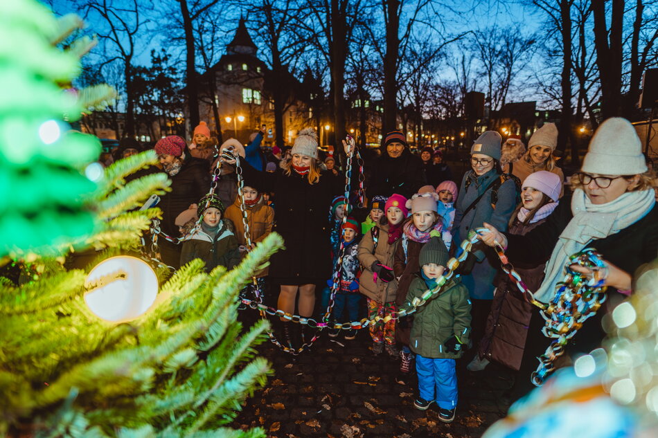 Na zdjęciu widać grupę dzieci i dorosłych otaczających świąteczną choinkę, udekorowaną lampkami, podczas wieczornego wydarzenia w parku. Ludzie trzymają kolorowe papierowe łańcuchy, a w tle migoczą światła i widać zarys budynków, co podkreśla radosny, świąteczny nastrój.