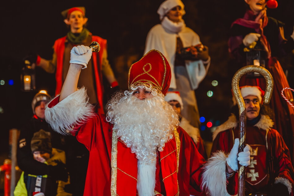 Na zdjęciu znajduje się postać przebrana za Świętego Mikołaja, trzymająca pastorał i dzwonek, ubrana w tradycyjny czerwony strój z białym futrem. W tle widoczne są inne postacie w świątecznych kostiumach, w tym elf i osoby z lampionami, co nadaje scenie uroczysty, bożonarodzeniowy klimat