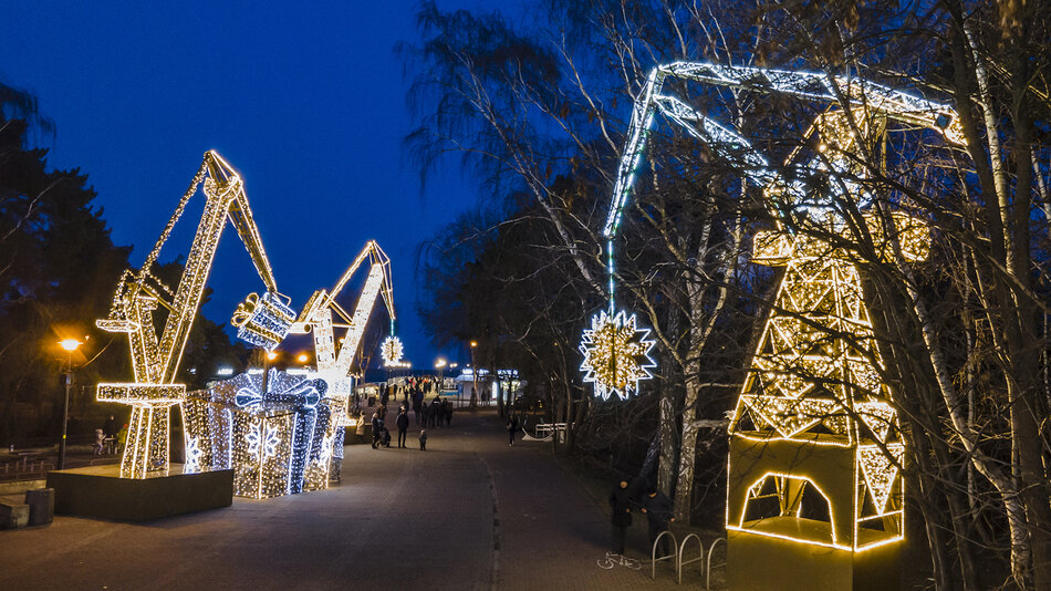 zdjęcie iluminacji zaprojektowanych w kształcie stoczniowych dźwigów i ogromnych prezentów, ustawionych wzdłuż szerokiego bulwaru, po którym chodzą ludzie. Całość otaczają drzewa. Zdjęcie wykonano wieczorem, po zachodzie słońca