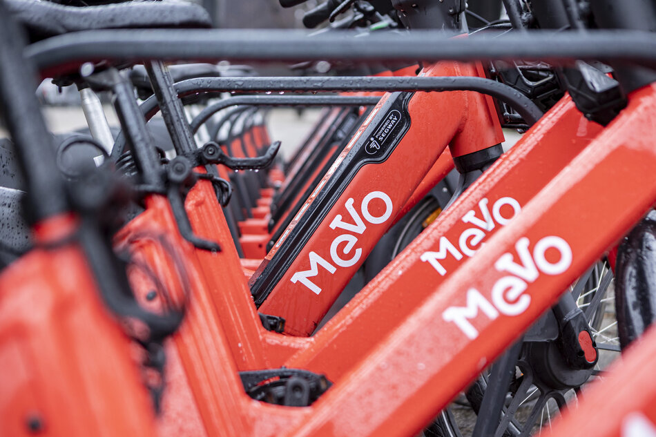 Zdjęcie przedstawia zbliżenie na rzędy czerwonych rowerów miejskich MEVO, ustawionych jeden obok drugiego. Charakterystyczne logo MEVO jest wyraźnie widoczne na ramach rowerów, które wyglądają na mokre, prawdopodobnie po deszczu.