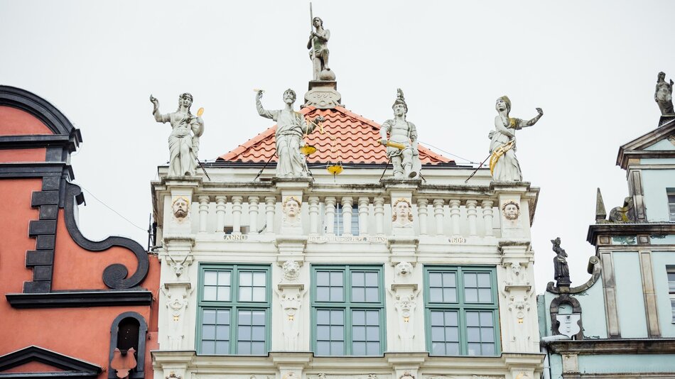 Na zdjęciu widać fragment fasady zabytkowego budynku o bogatej architekturze. Budowla posiada klasyczne, zdobione detale charakterystyczne dla stylu manieryzmu lub baroku. Na górze budynku znajduje się kilka rzeźb przedstawiających postacie, prawdopodobnie alegoryczne. Są one umieszczone na gzymsach i symetrycznie rozmieszczone. Centralnie na szczycie znajduje się dodatkowa figura trzymająca długi przedmiot, przypominający broń lub narzędzie. Elewacja budynku jest bogato zdobiona pilastrami, kolumnami i ornamentami. Na ścianie widać również twarze wyrzeźbione w kamieniu, które mogą symbolizować różne wartości lub odnosić się do historii budynku. Pod rzeźbami znajduje się czerwony dach z ozdobnymi elementami w postaci złotych akcentów. Budynek jest otoczony przez inne historyczne kamienice - po lewej stronie widać fragment czerwonej fasady z czarnymi detalami, natomiast po prawej stronie jest błękitna kamienica ze zdobionym szczytem. Całość tworzy kompozycję typową dla europejskiego historycznego rynku miejskiego, prawdopodobnie zlokalizowanego w jednym z miast o bogatym dziedzictwie architektonicznym, jak Gdańsk czy inne miasta hanzeatyckie.