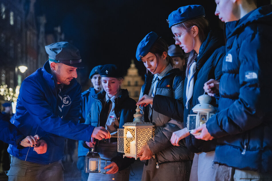 Zdjęcie przedstawia grupę młodych ludzi w mundurach harcerskich i ciepłych kurtkach, którzy wspólnie trzymają i przekazują lampiony ze światłem. Scena odbywa się w nocy na tle miasta, a atmosfera jest skupiona i symboliczna, sugerując uroczystość lub ważne wydarzenie związane z tradycją przekazywania światła pokoju.