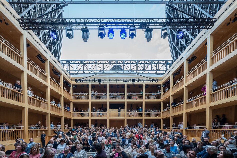 Na zdjęciu przedstawiono wnętrze Gdańskiego Teatru Szekspirowskiego z widownią pełną ludzi, którzy czekają na rozpoczęcie przedstawienia lub wydarzenia. Charakterystyczna drewniana konstrukcja galerii oraz otwierany dach nadają przestrzeni wyjątkowy, klasyczny wygląd inspirowany dawnymi teatrami elżbietańskimi