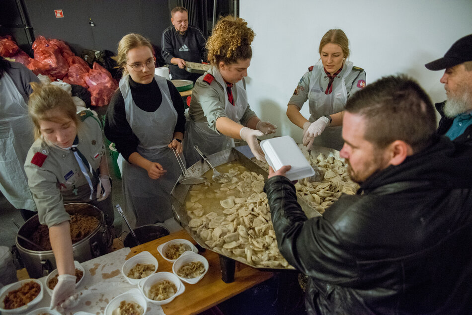 Na zdjęciu widać grupę młodych ludzi w harcerskich mundurach i fartuchach, którzy rozdają ciepły posiłek potrzebującym osobom. Duże naczynie z pierogami i miski z dodatkami stoją na stole, a wolontariusze starannie obsługują zgromadzone osoby, przekazując jedzenie w plastikowych pojemnikach