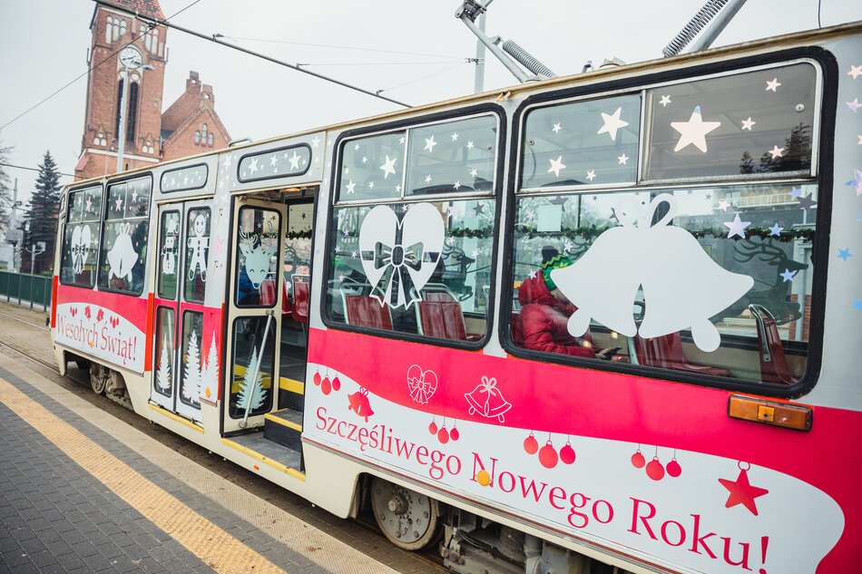 Na zdjęciu znajduje się świątecznie udekorowany tramwaj z napisami Wesołych Świąt! oraz Szczęśliwego Nowego Roku!, ozdobiony białymi motywami świątecznymi, takimi jak dzwonki, serca i gwiazdy. W tle widoczny jest budynek z zegarem, który przypomina kościół, oraz fragment torowiska