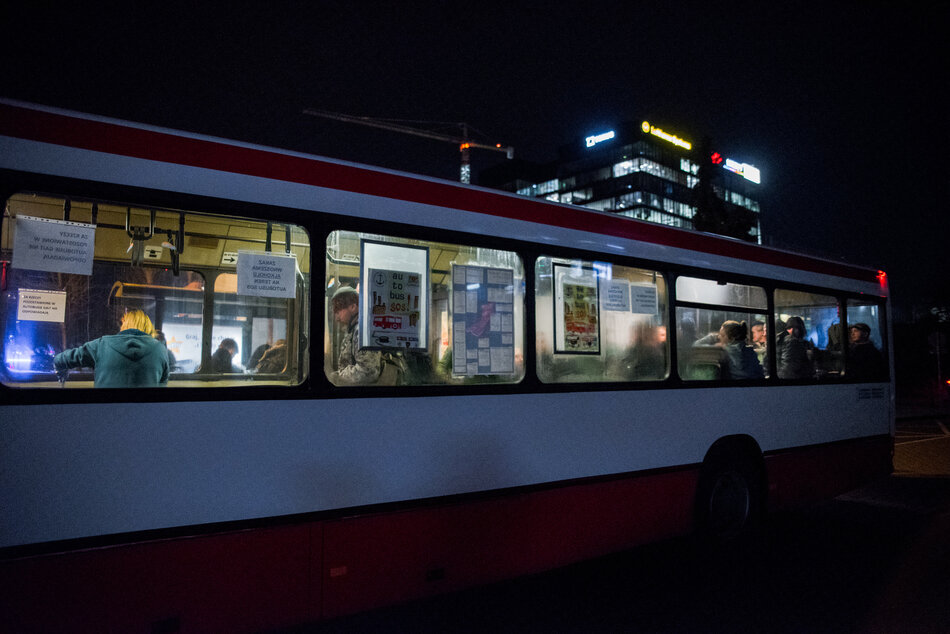 Zaparkowany autobus na zimowej ciemnej ulicy. Szyby są zaparowane, ale widać że w środku znajdują się osoby.