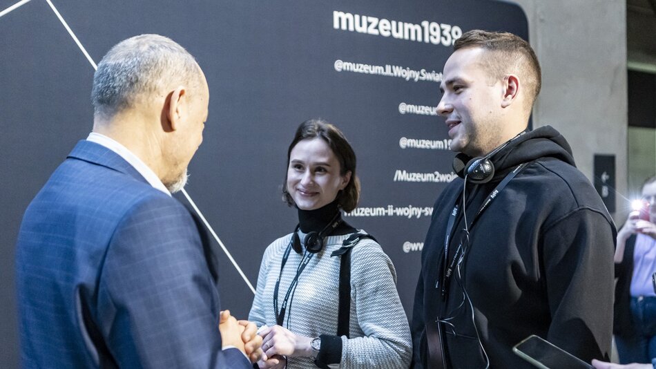 Na zdjęciu widzimy trzy osoby w interakcji, prawdopodobnie podczas wydarzenia w Muzeum II Wojny Światowej w Gdańsku, co sugeruje widoczne w tle napisy muzeum1939 i powiązane linki do mediów społecznościowych. Po lewej stronie znajduje się mężczyzna w średnim wieku, widoczny od tyłu, ubrany w elegancki granatowy garnitur. W centrum kadru stoi młoda kobieta z krótkimi, brązowymi włosami, ubrana w jasny sweter i czarny golf. Na szyi ma zawieszone słuchawki oraz identyfikator. Uśmiecha się lekko, patrząc na mężczyznę w garniturze. Po prawej stronie jest młody mężczyzna w czarnej bluzie, również z identyfikatorem i słuchawkami na szyi, który także wygląda na zaangażowanego w rozmowę. W tle widać fragmenty osób, w tym jedną robiącą zdjęcie. Atmosfera wydaje się być formalna, ale przyjazna.