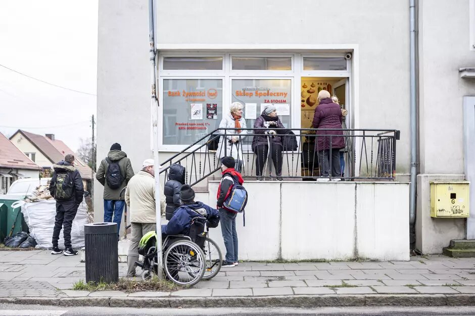 Kolejka osób stojących pod sklepem. W kolejce jest osoba na wózku inwalidzkim. 