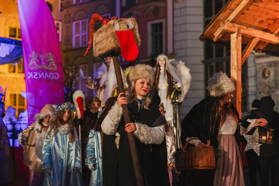 Zdjęcie przedstawia grupę ludzi ubranych w tradycyjne i fantazyjne stroje podczas świątecznego wydarzenia w Gdańsku, co sugeruje charakter jarmarku bożonarodzeniowego lub kolędniczego pochodu. Na pierwszym planie widoczna jest kobieta w futrzanej czapce, trzymająca drewniany kij z ozdobną figurą przypominającą smoka lub potwora, a wokół niej znajdują się osoby w anielskich i historycznych kostiumach